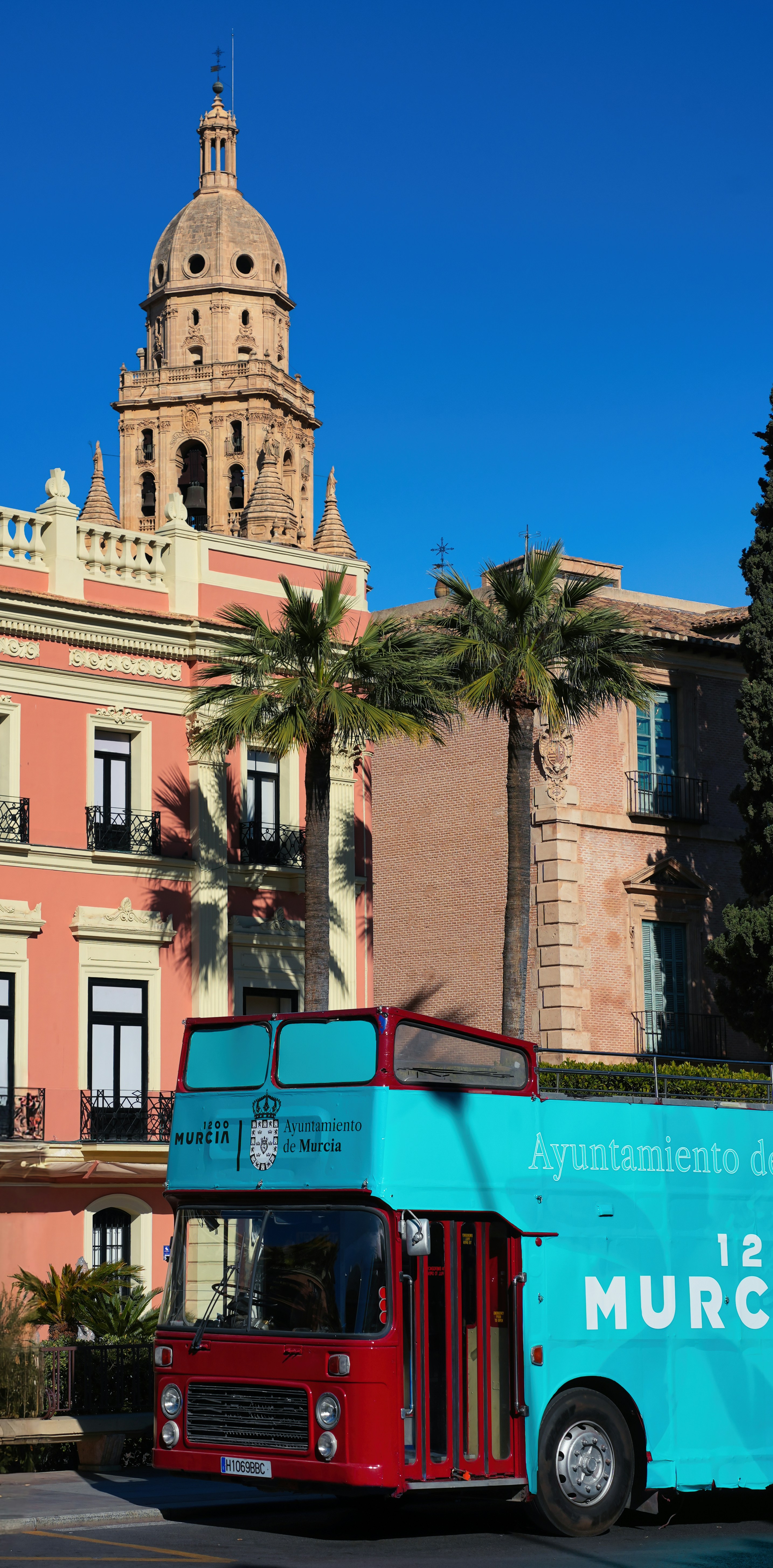 Open-top tour bus passing through Murcia, with the cathedral tower and palm-lined historic buildings in the background.