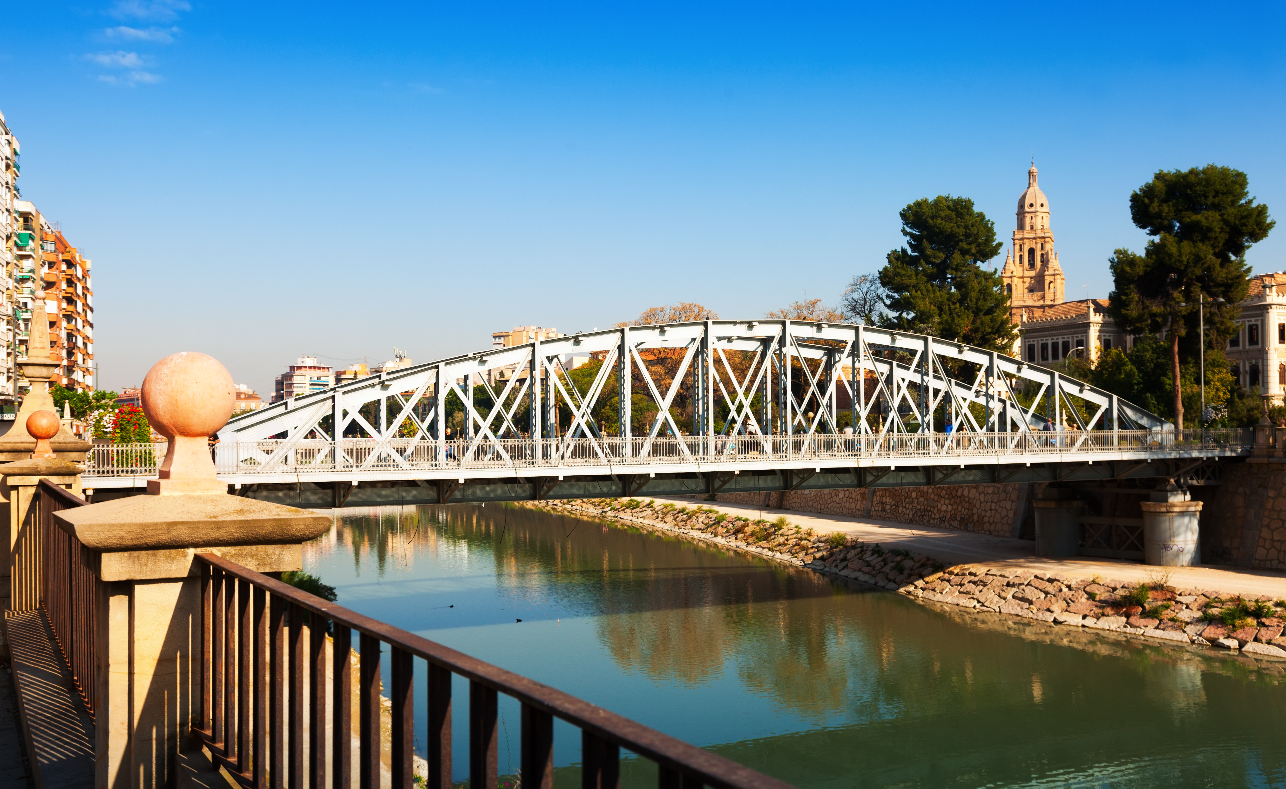 Segura River in Murcia with a pedestrian bridge and cathedral tower in the distance, near the Floridablanca Gardens and El Carmen area