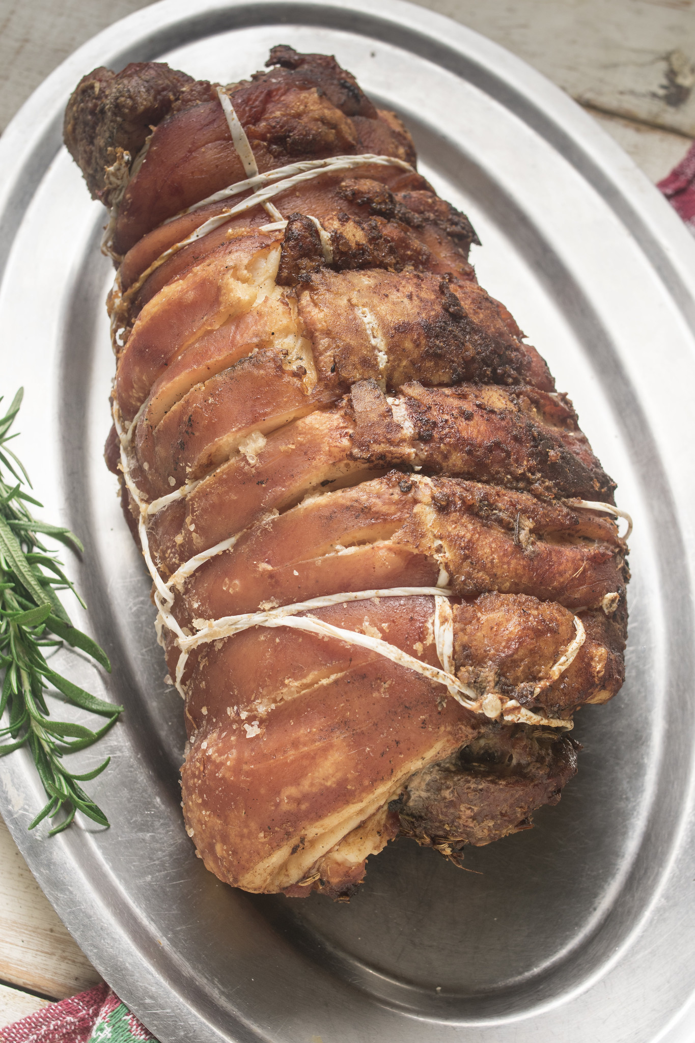 Slow-roasted porchetta with golden crispy crackling, tied with string and served on a metal platter with rosemary.