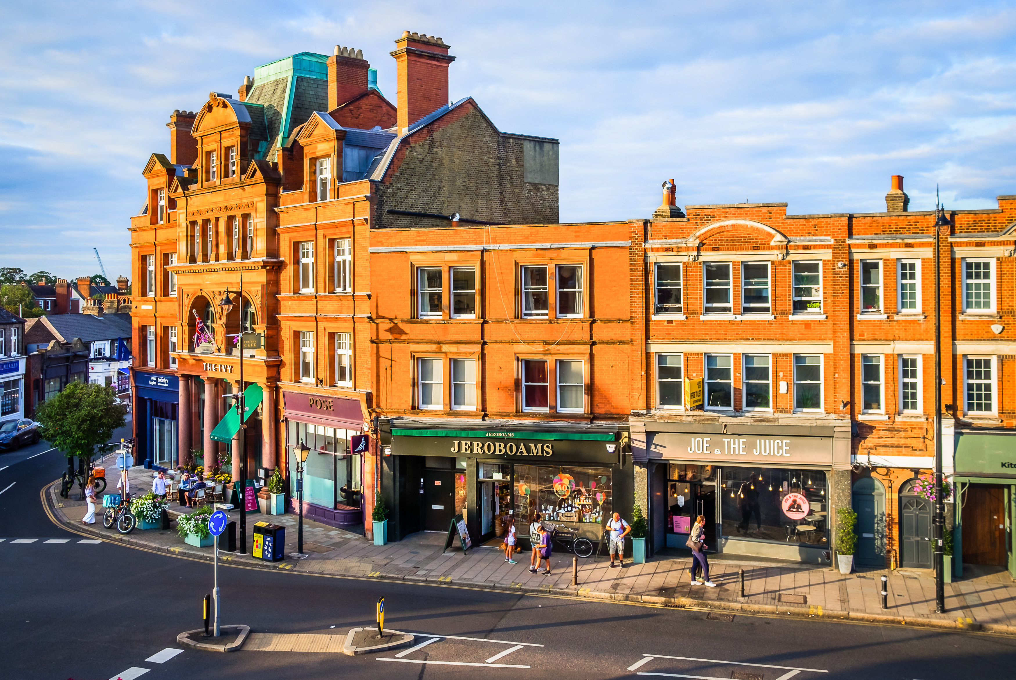 Wimbledon Village street with red-brick buildings, shops, cafés and hotel frontage in warm evening light.